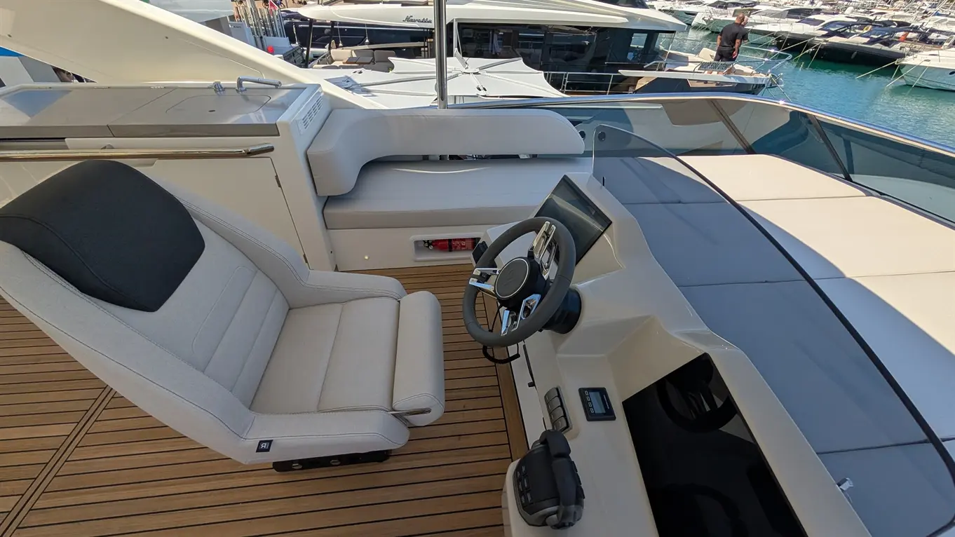 Luxury yacht cockpit featuring a beige captain's chair, steering wheel, and navigation screens, surrounded by docked boats.
